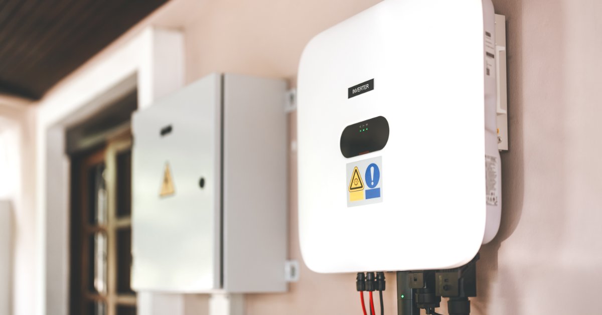 A wall-mounted white inverter and gray electrical cabinet with warning labels and cables attached outside a building.