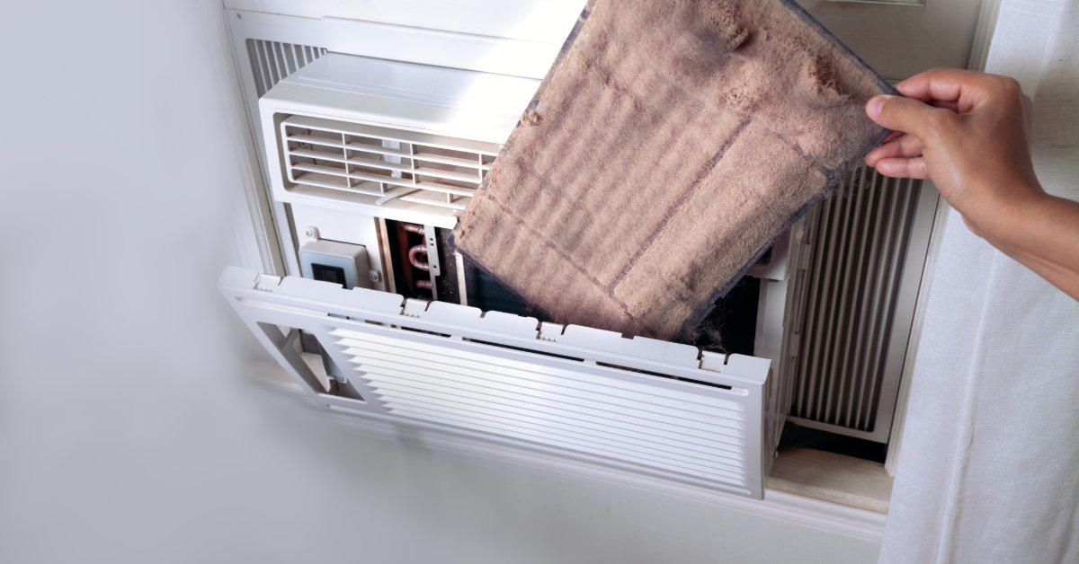 A hand pulling a dusty filter from a white window air conditioner mounted beside a curtain and wall vent.