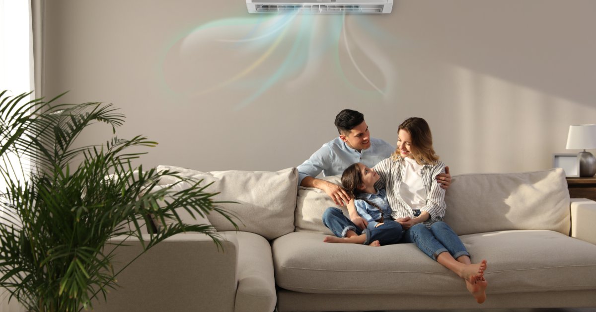Two adults and a child sit on a beige couch below a wall air conditioner, with a green plant near the window.