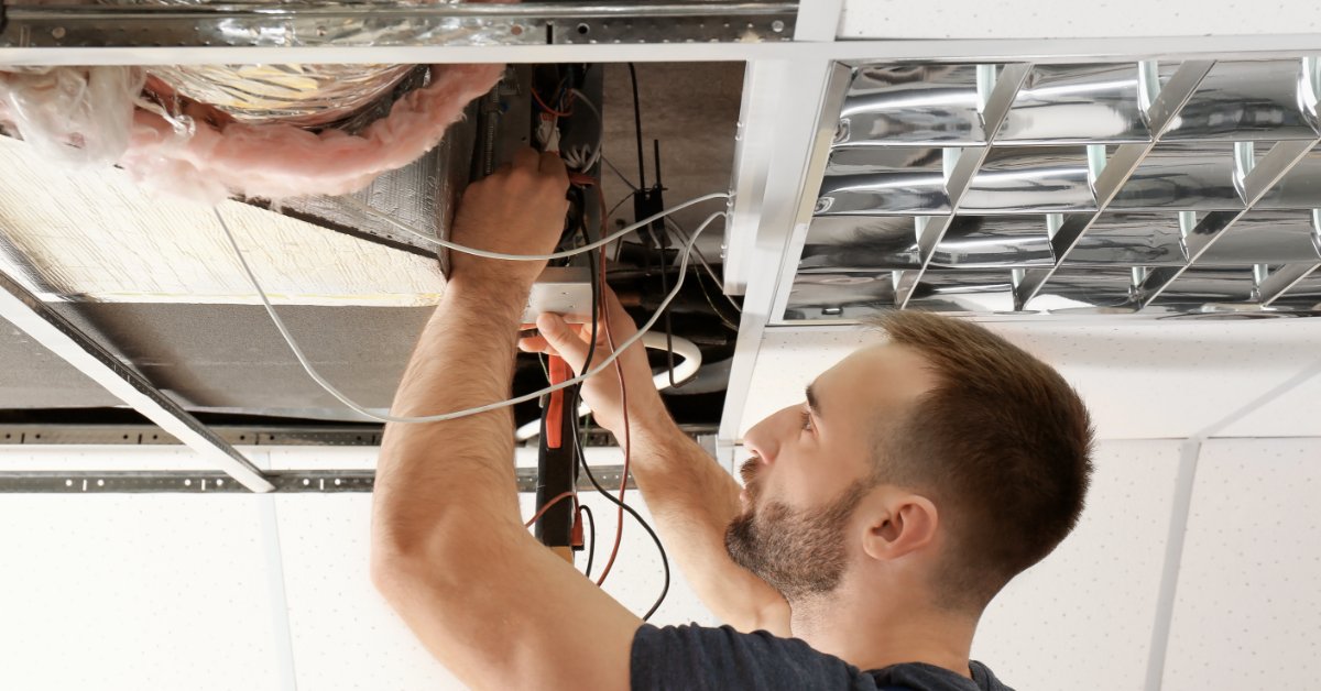 A close-up of a person holding multimeter probes near wiring inside an open ceiling-mounted air conditioning unit.