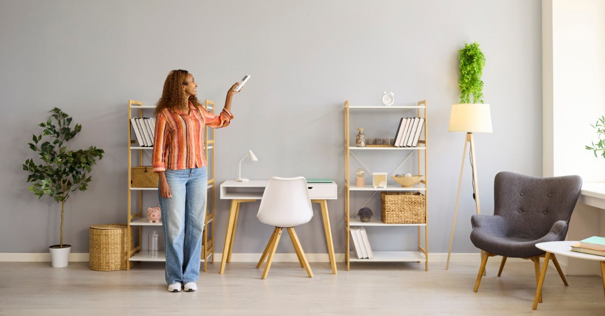  A woman holds a remote toward a wall-mounted air conditioner in a living room with shelves and seating.