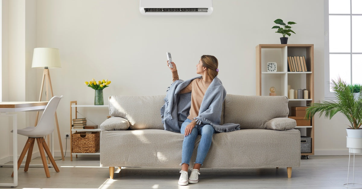 A woman sitting on a sofa holds a remote in her right hand and points it at an air conditioning system on the wall.