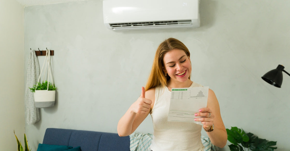 A woman standing in front of a wall-mounted mini split system gives a thumbs up as she looks at a paper with a graph on it.