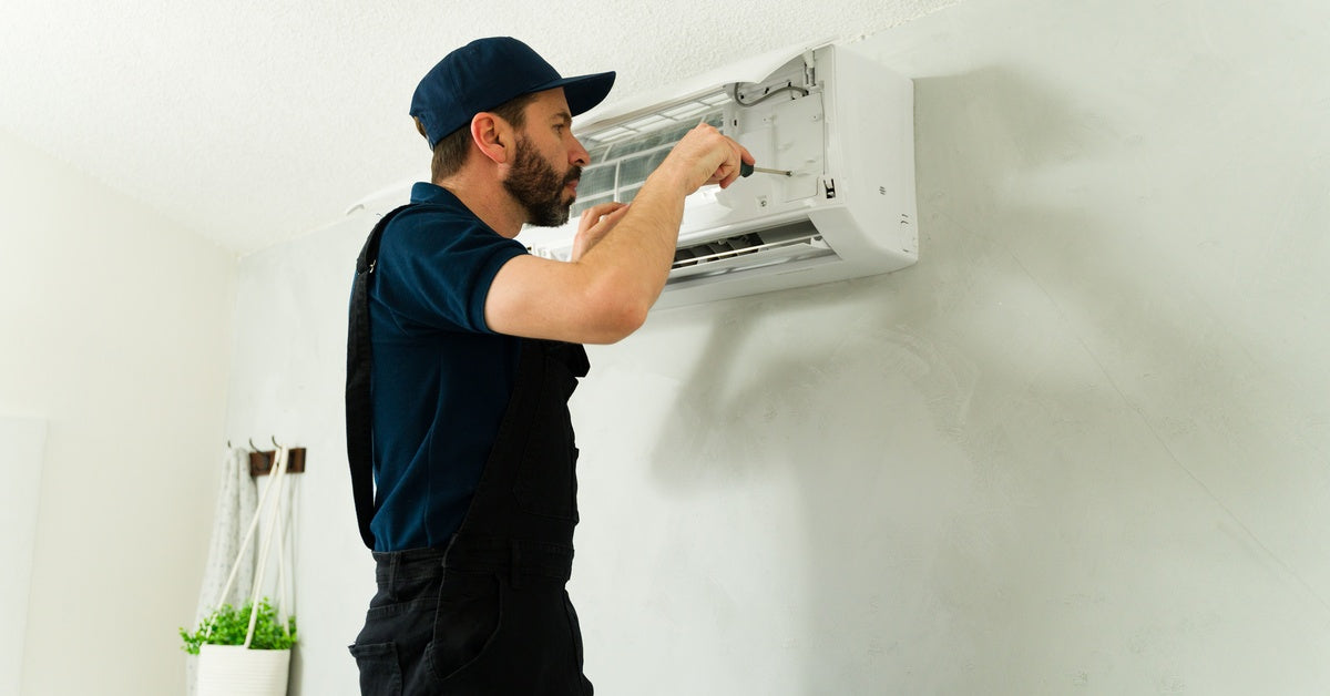 A person wearing a blue shirt and matching cap and black overalls places a screwdriver in a wall-mounted AC unit.