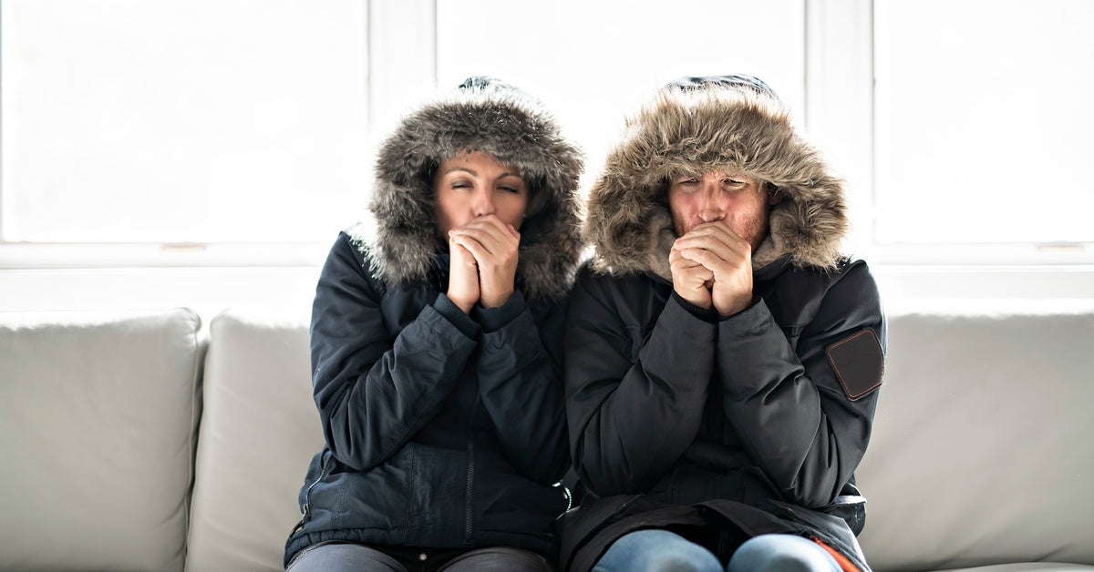 A couple sits on the sofa in their home. They’re wearing heavy winter coats and blowing on their hands to keep warm.