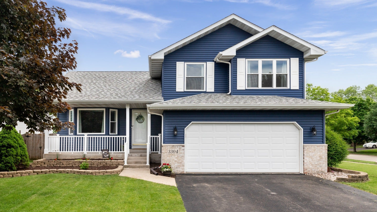 A blue two-story suburban home with white trim, a front porch, and a garage, surrounded by green lawn, trees, and a landscaped yard.