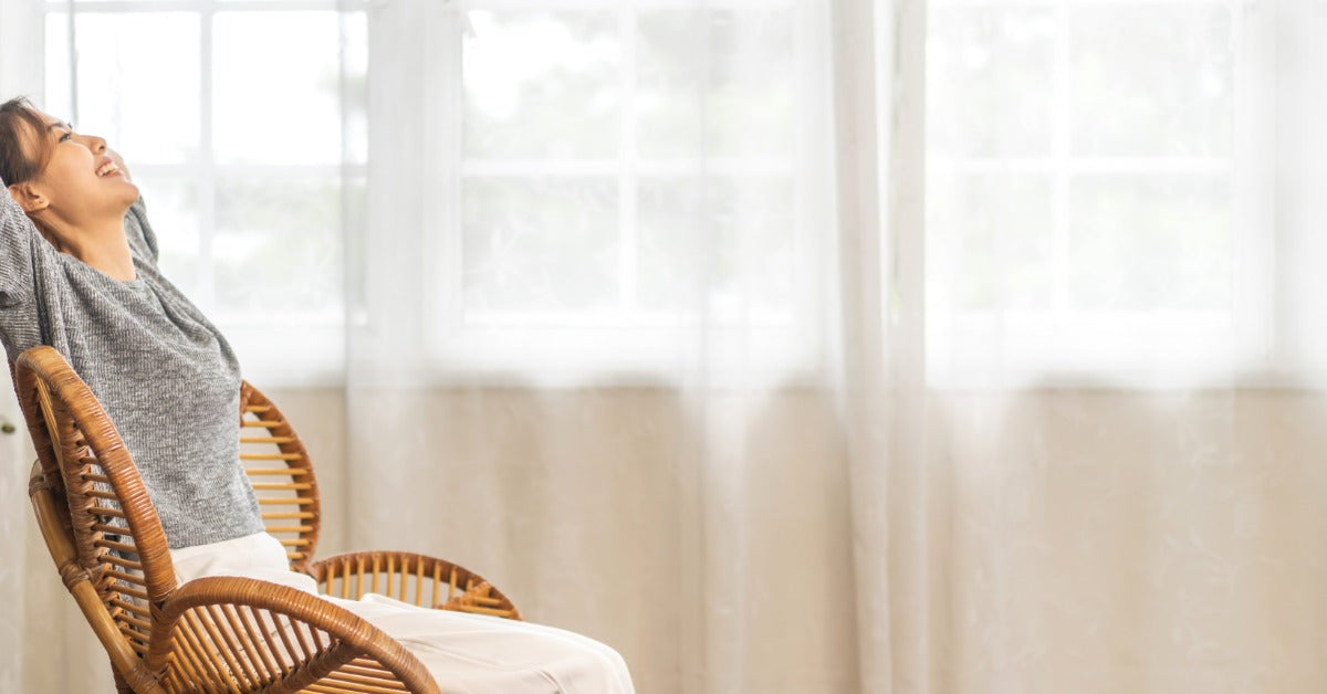 Woman sitting relaxed in a rattan chair by a bright window, smiling and enjoying perfect air balance in her home.