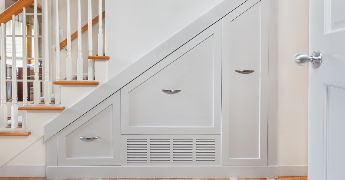 Built-in white cabinets with a concealed mini-split vent beneath a wooden staircase in a bright home interior.